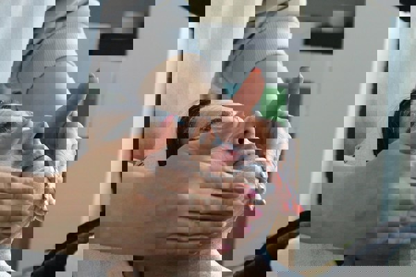 Person receiving a facial treatment while lying down, wearing a white headband, as a practitioner applies a cream or mask to their face. Skincare products are visible on shelves in the background.